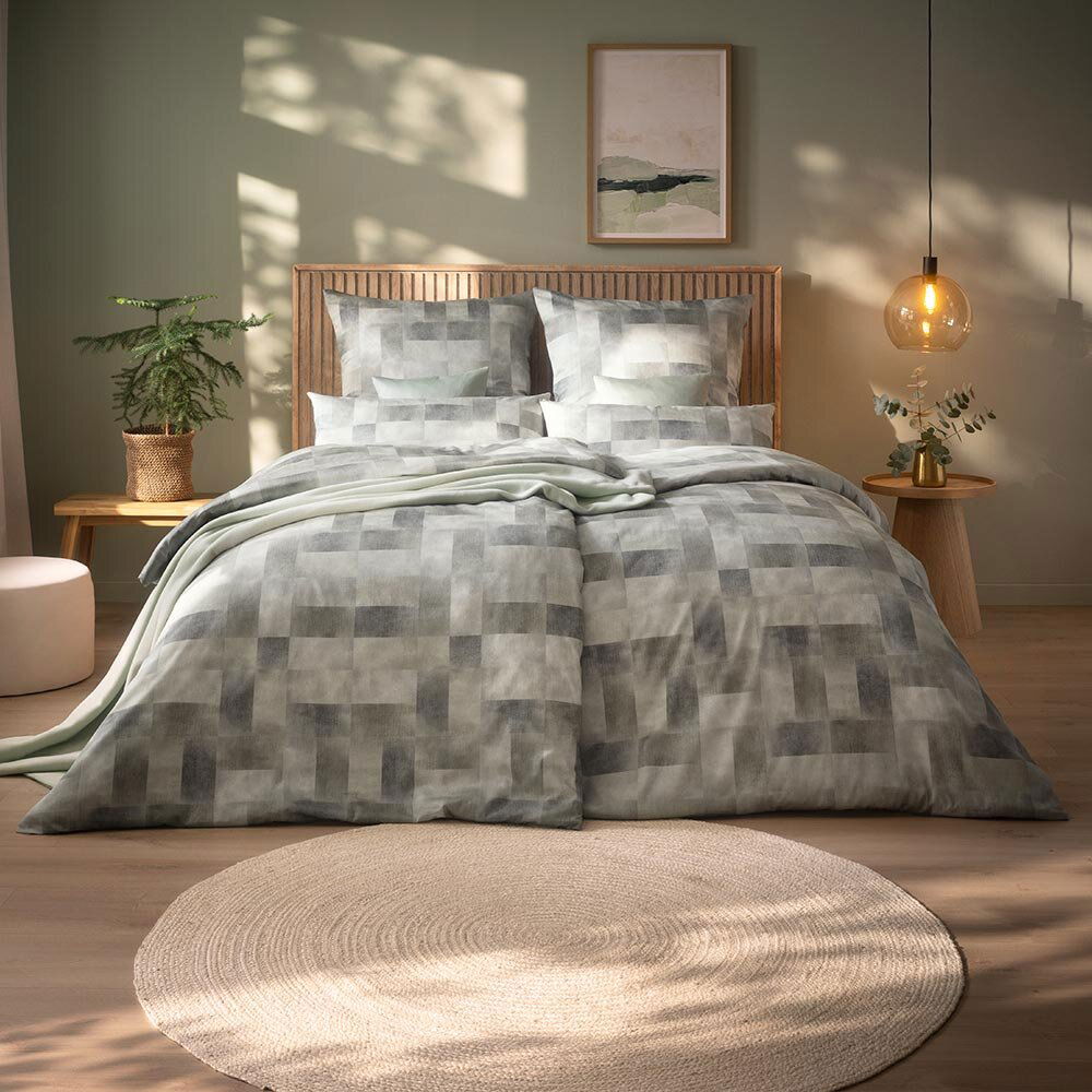 Bett mit gemustertem Bettbezug in einem modernen Schlafzimmer, frontal fotografiert. Im Hintergrund sind ein Holz-Kopfteil, ein Pflanzenarrangement und eine hängende Lampe zu sehen.