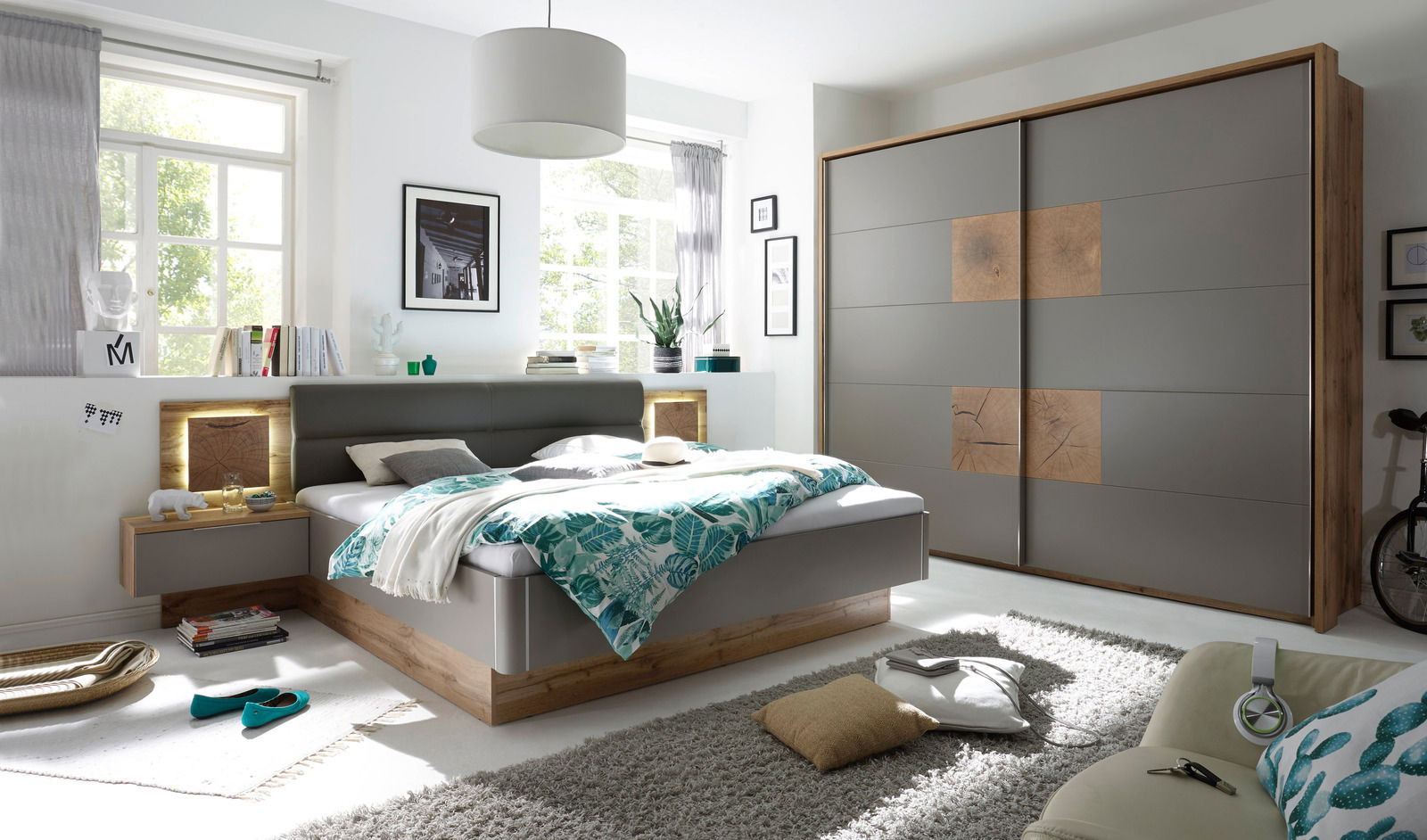 Modernes Schlafzimmer mit einem grauen Schwebetürenschrank und einem Bett mit Holz- und Grauelementen, seitliche Perspektive.