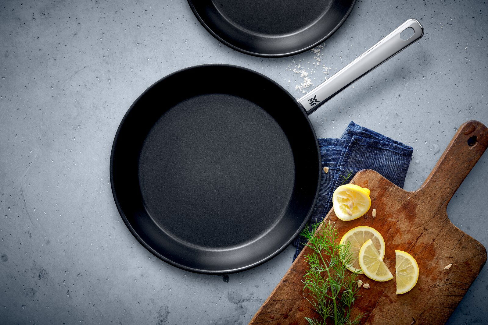 WMF Pfannenset 2-tlg. NORDIC PROFI Pfannenset Nordics, bestehend aus zwei schwarzen Pfannen in den Größen 24 cm und 28 cm, von oben fotografiert. Daneben ein Holzbrett mit Zitronenscheiben und Kräutern.