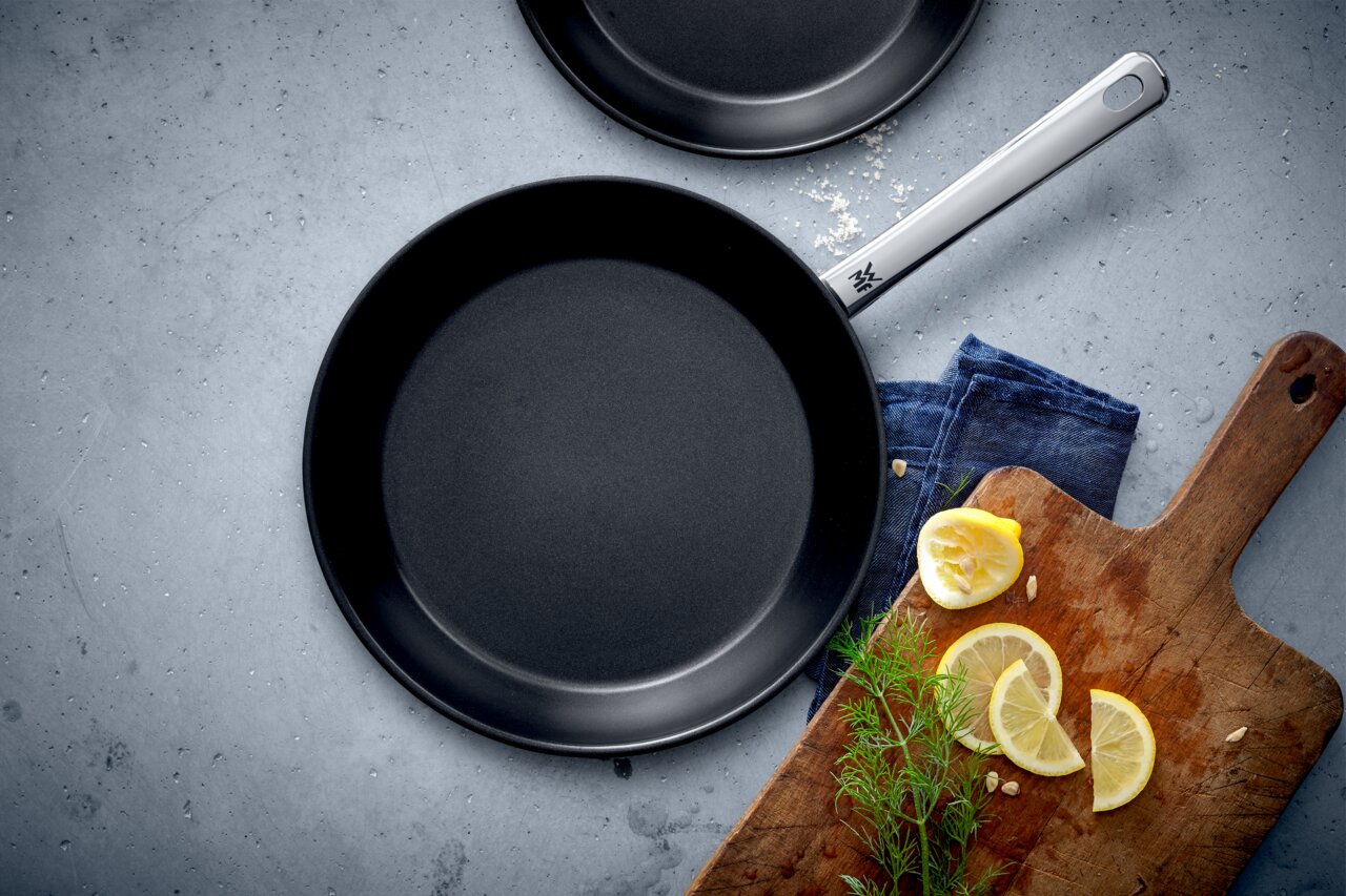 WMF Pfannenset 2-tlg. NORDIC PROFI Pfannenset Nordics, bestehend aus zwei schwarzen Pfannen in den Größen 24 cm und 28 cm, von oben fotografiert. Daneben ein Holzbrett mit Zitronenscheiben und Kräutern.