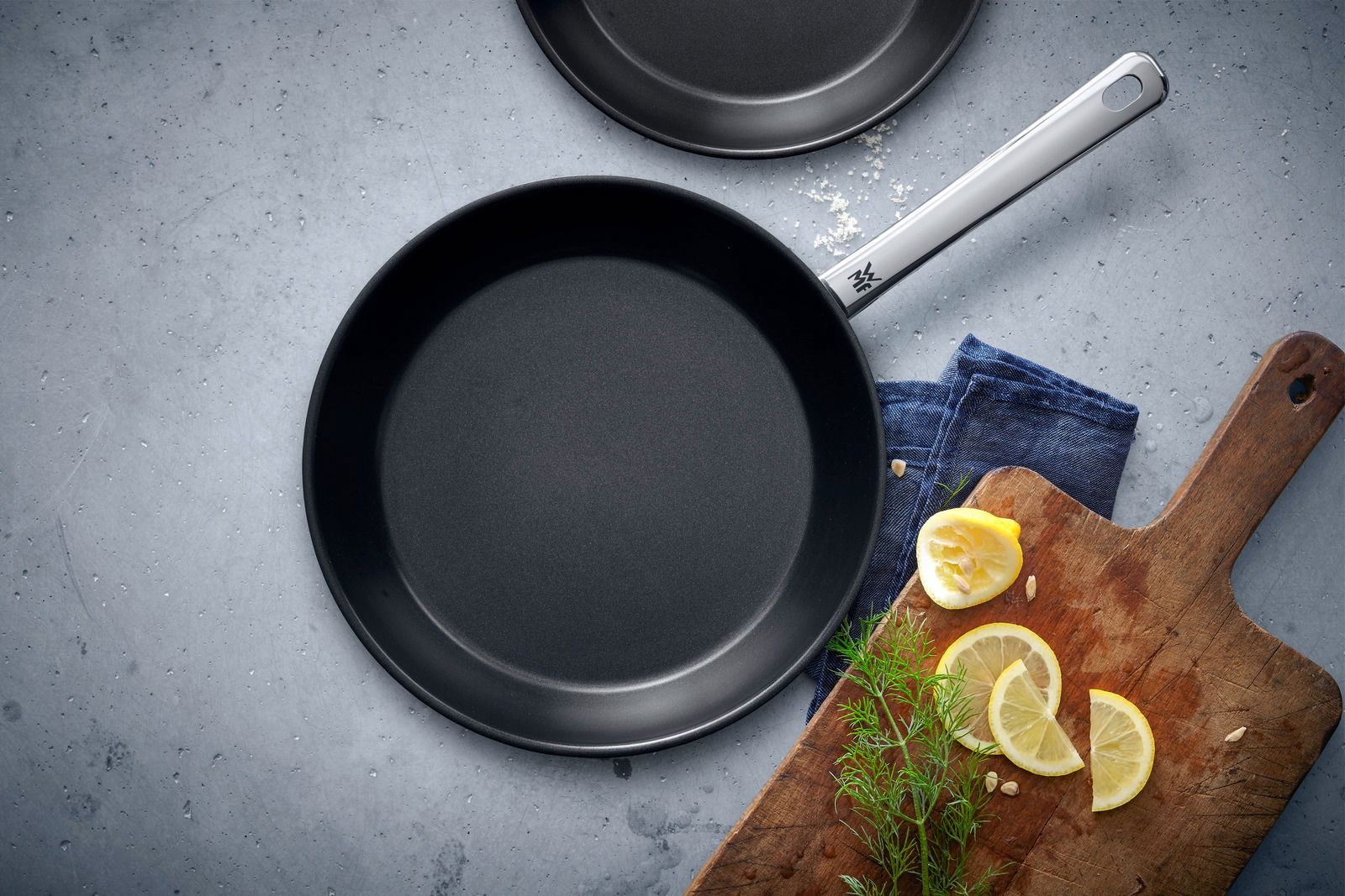WMF Pfannenset 2-tlg. NORDIC PROFI Pfannenset Nordics, bestehend aus zwei schwarzen Pfannen in den Größen 24 cm und 28 cm, von oben fotografiert. Daneben ein Holzbrett mit Zitronenscheiben und Kräutern.