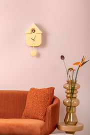 Gelbe Wanduhr in Form eines Vogelhauses mit Pendel, an einer rosa Wand über einem orangefarbenen Sofa und neben einer Vase mit Blumen. Frontalansicht.