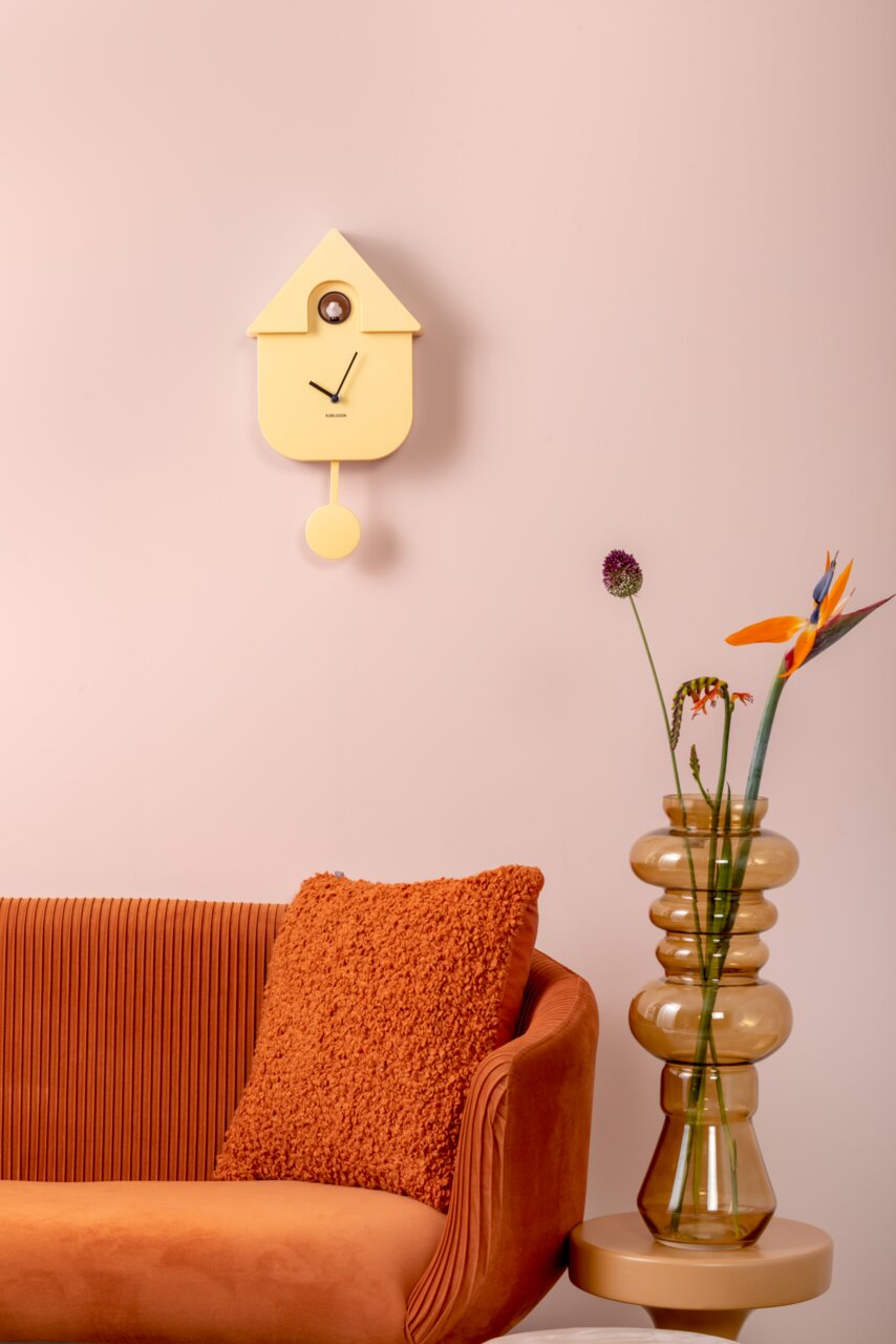 Gelbe Wanduhr in Form eines Vogelhauses mit Pendel, an einer rosa Wand über einem orangefarbenen Sofa und neben einer Vase mit Blumen. Frontalansicht.