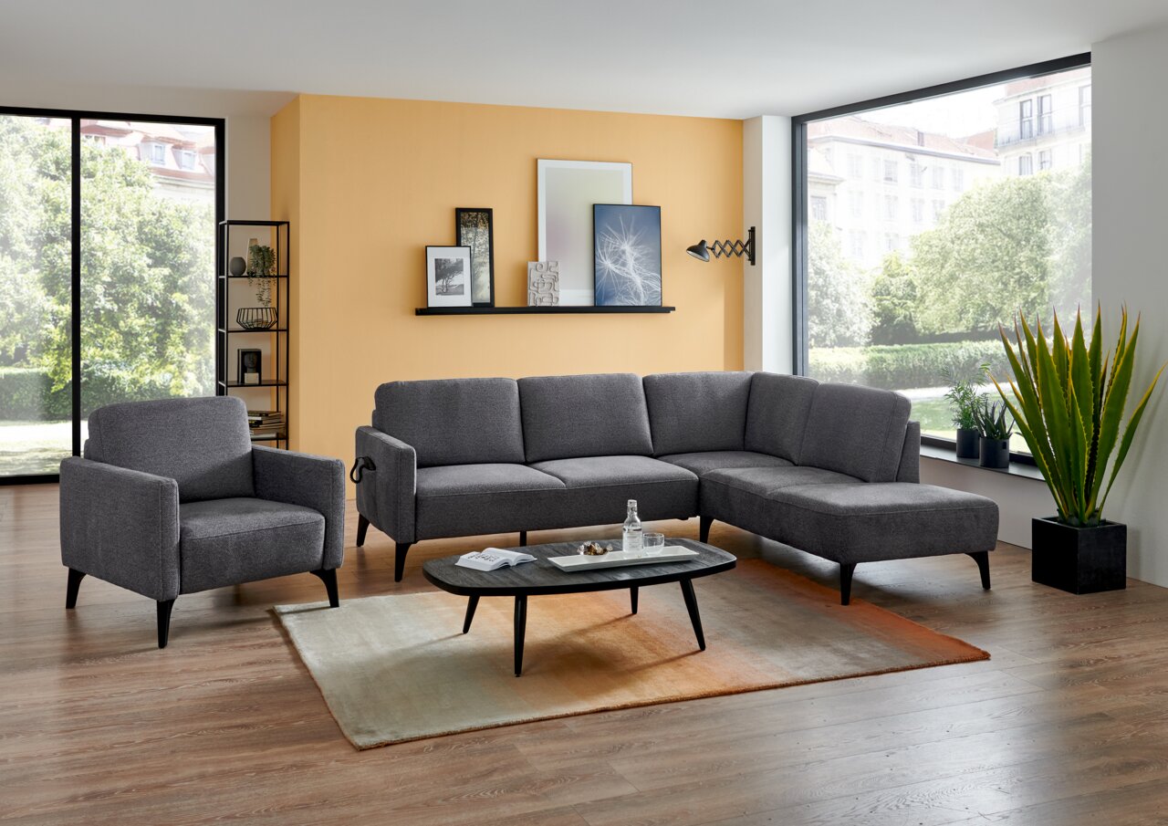 Modernes Wohnzimmer mit grauem Stoff-Ecksofa mit Recamiere rechts und passendem Sessel, aufgenommen in frontaler leicht schräger Perspektive; davor schwarzer Couchtisch auf Teppich, große Fenster und orangefarbene Akzentwand im Hintergrund.