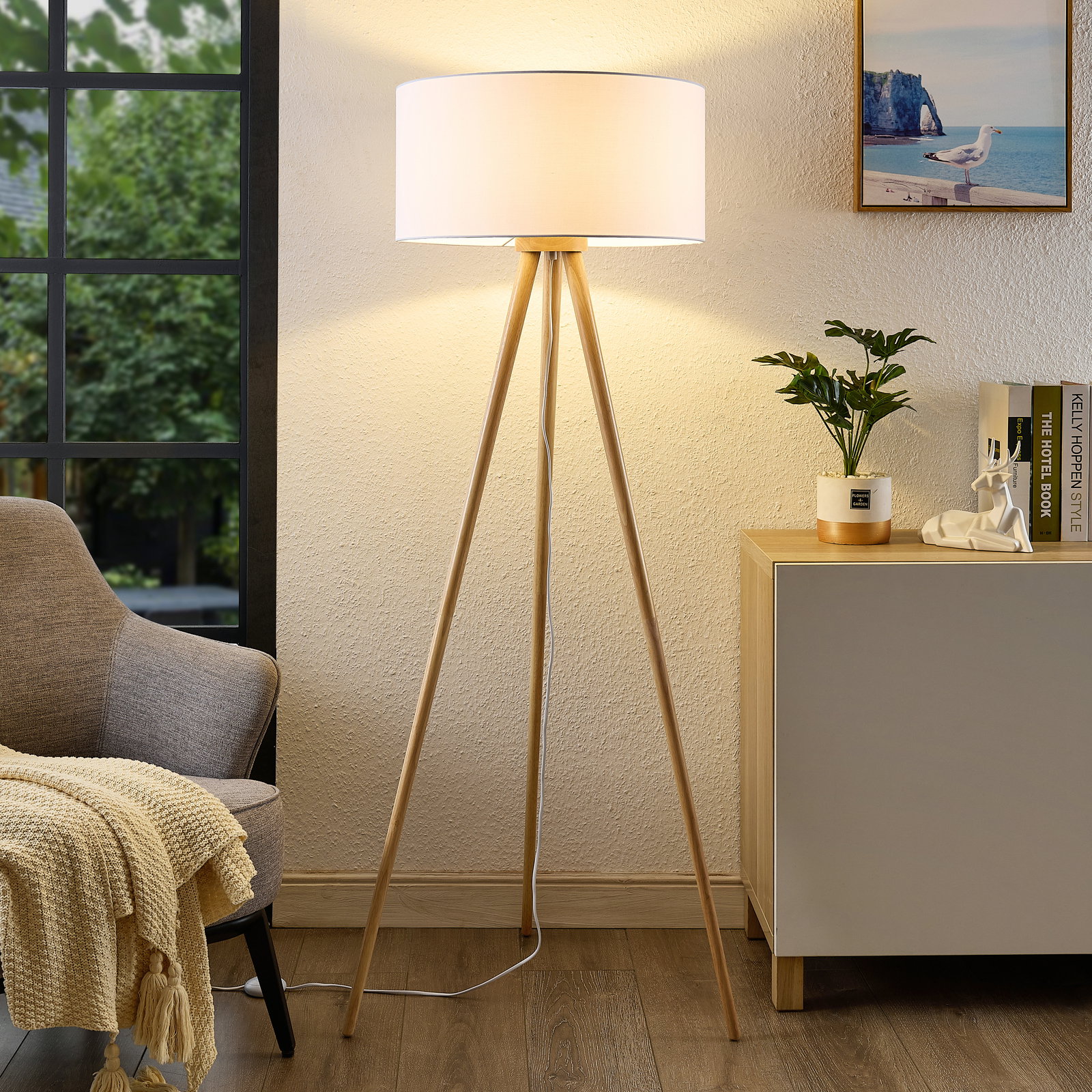 Moderne Stehleuchte mit dreibeinigem Holzgestell und weißem Lampenschirm, frontal im Wohnzimmer platziert.