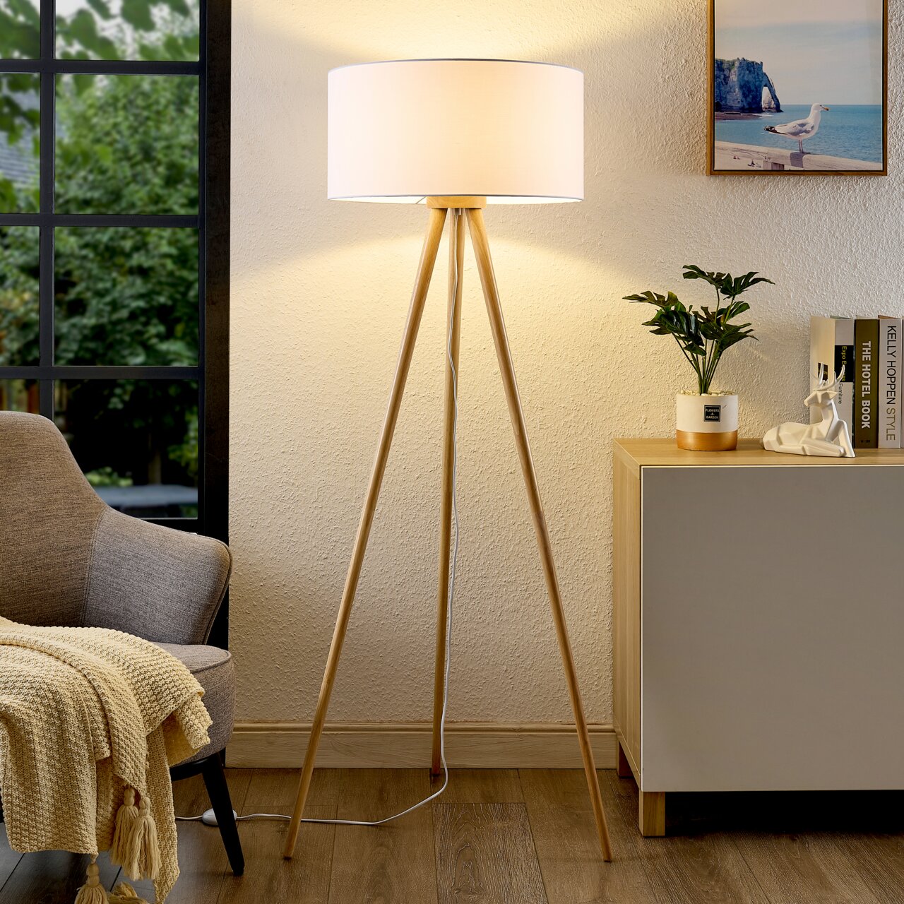 Moderne Stehleuchte mit dreibeinigem Holzgestell und weißem Lampenschirm, frontal im Wohnzimmer platziert.