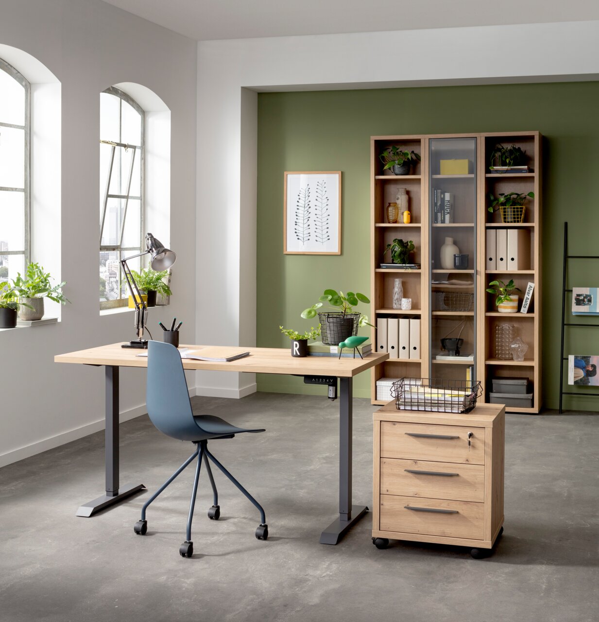 Moderner Arbeitsplatz mit elektrischem Schreibtischgestell und Tischplatte in einem hellen Raum mit grüner Akzentwand, Pflanzen und Regalen. Perspektive von vorne.