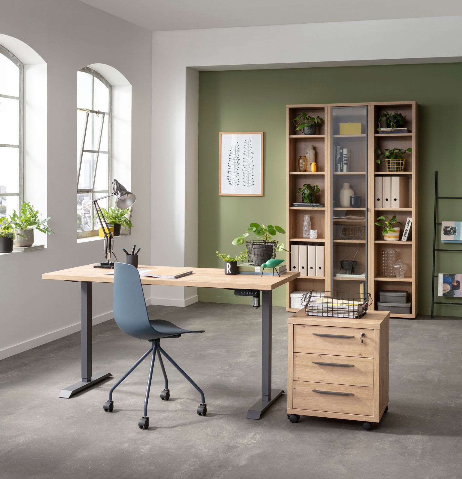 Moderner Arbeitsplatz mit elektrischem Schreibtischgestell und Tischplatte in einem hellen Raum mit grüner Akzentwand, Pflanzen und Regalen. Perspektive von vorne.