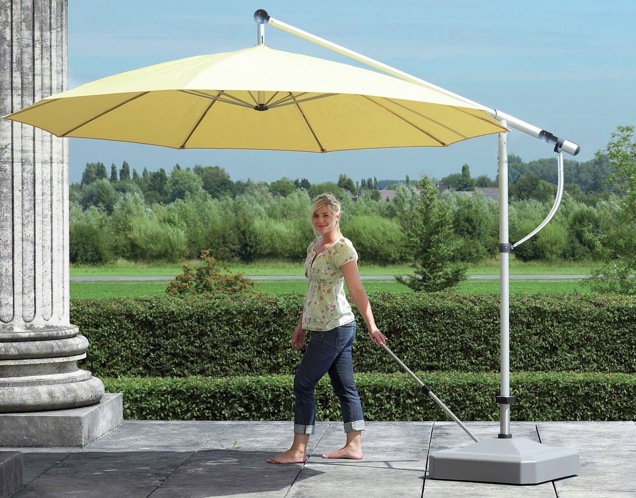 Frau steht neben einem gelben Sonnenschirm auf einer Terrasse mit Blick auf einen Garten, seitliche Perspektive