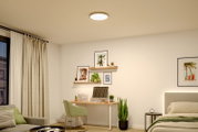 Runde, flache LED-Deckenleuchte in Weiß mit warmem Licht, montiert an der Zimmerdecke in einem modern eingerichteten Schlafzimmer/Homeoffice; Ansicht leicht von unten (Untersicht).