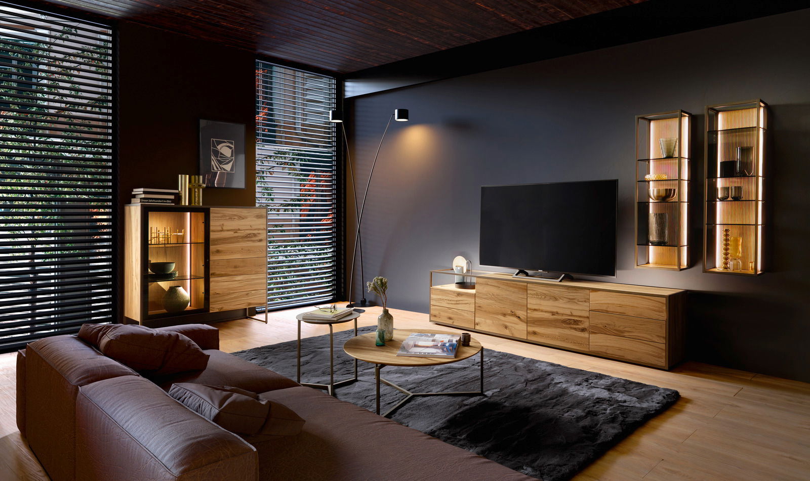Modernes Wohnzimmer mit einem Highboard aus Holz, einem großen Fernseher auf einem Sideboard und zwei beleuchteten Hängeregalen an der Wand. Perspektive von der Seite.