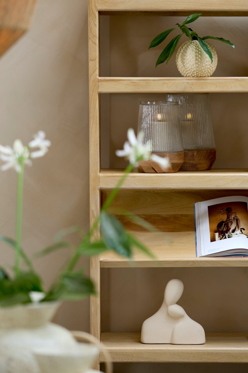 Holzregal mit dekorativen Elementen: Oben eine runde, strukturierte Vase mit grünen Blättern, daneben zwei geriffelte Glaslichter mit Kerzen. Unten eine abstrakte Skulptur und ein aufgeschlagenes Buch. Perspektive von vorne.