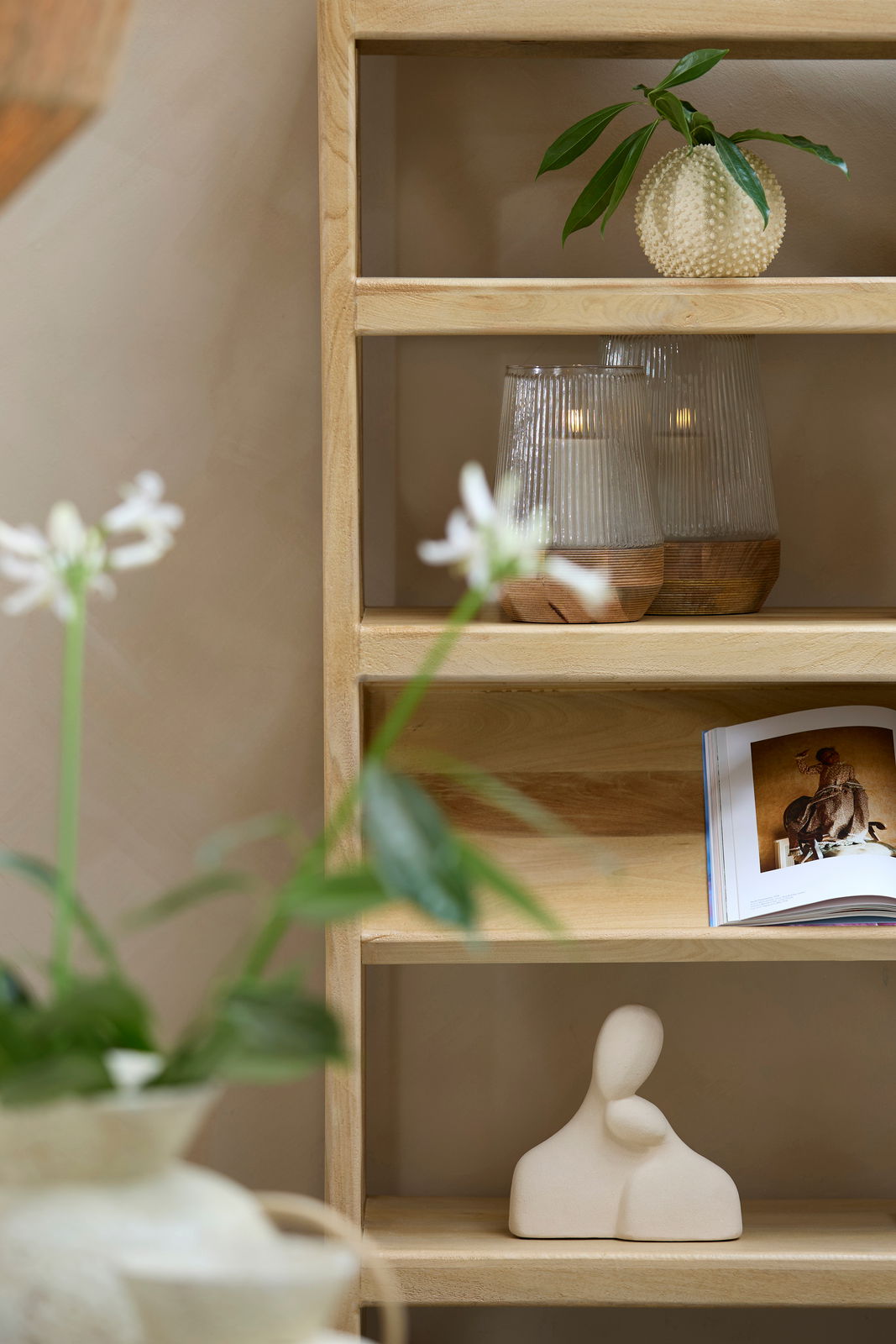 Holzregal mit dekorativen Elementen: Oben eine runde, strukturierte Vase mit grünen Blättern, daneben zwei geriffelte Glaslichter mit Kerzen. Unten eine abstrakte Skulptur und ein aufgeschlagenes Buch. Perspektive von vorne.