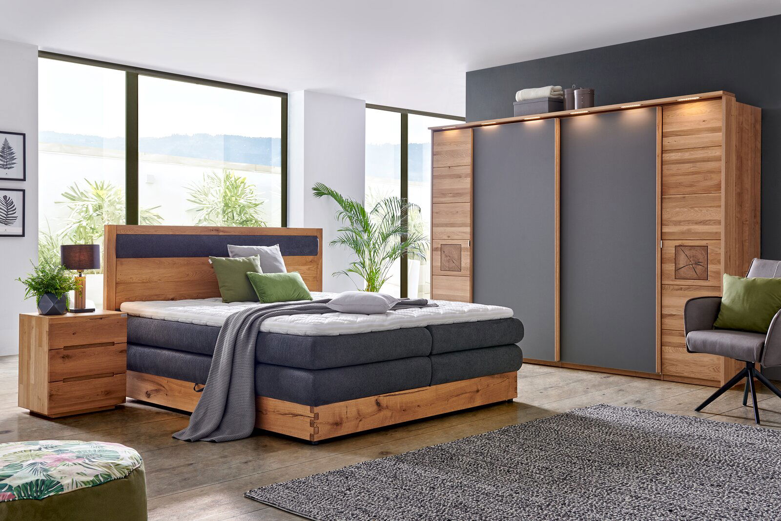 Moderne Schlafzimmeransicht mit einem Schwebetürenschrank in Holzoptik und grauen Türen, einem gemütlichen Bett mit Holzrahmen und grauer Polsterung, sowie einem Nachttisch. Die Perspektive zeigt das Zimmer von vorne mit großen Fenstern im Hintergrund.