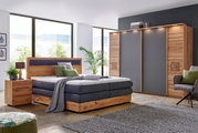 Moderne Schlafzimmeransicht mit einem Schwebetürenschrank in Holzoptik und grauen Türen, einem gemütlichen Bett mit Holzrahmen und grauer Polsterung, sowie einem Nachttisch. Die Perspektive zeigt das Zimmer von vorne mit großen Fenstern im Hintergrund.