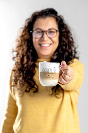 Frau in gelbem Pullover hält eine Tasse mit der Aufschrift 'SMILE' in der Hand, gefüllt mit einem Getränk. Frontalansicht.