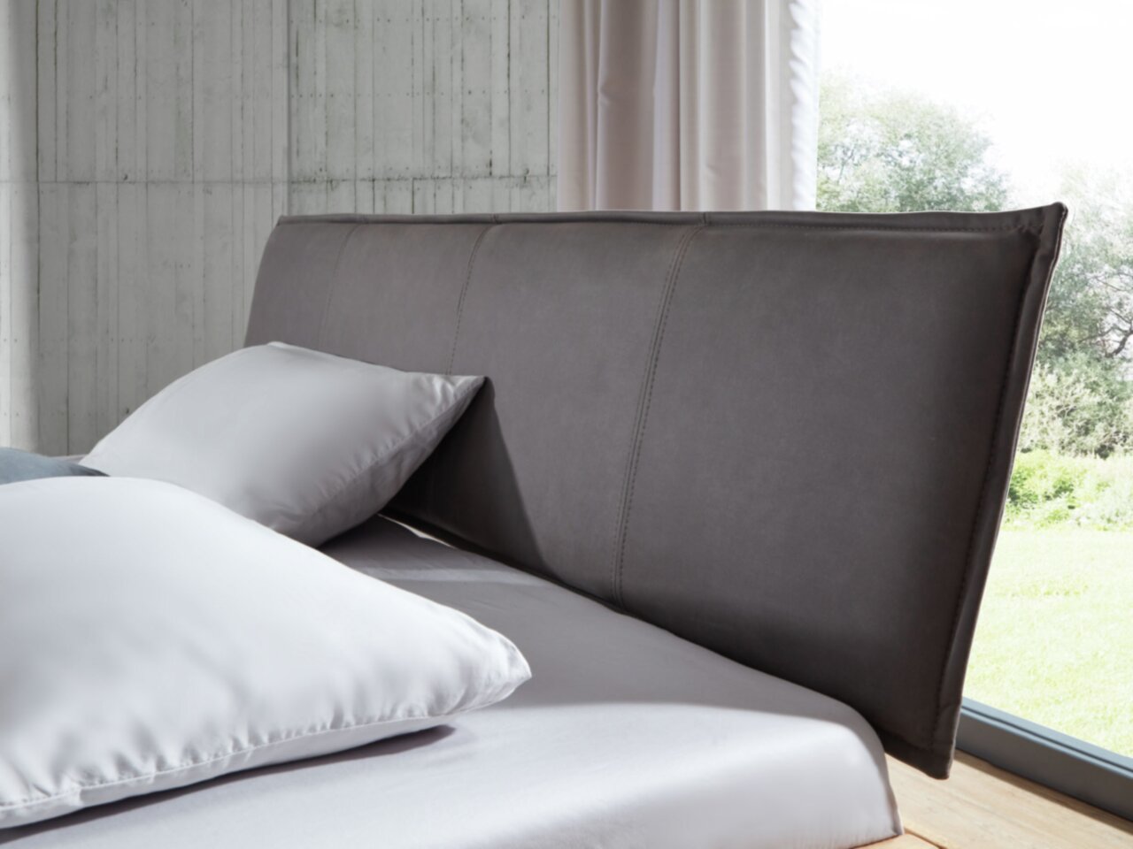 Nahaufnahme eines Balkenbetts mit grauem Kopfteil und weißen Kissen, seitliche Perspektive.