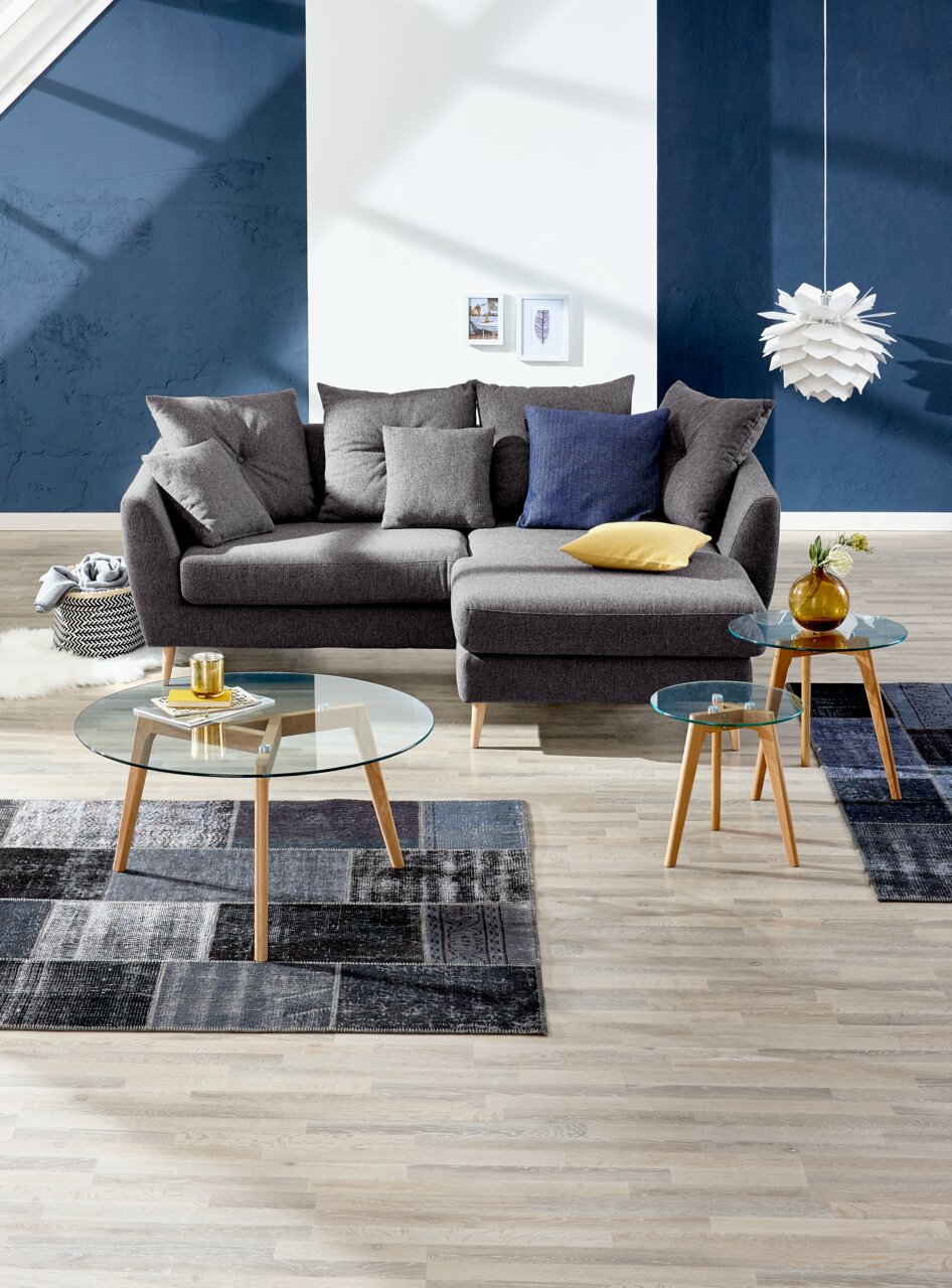 Graue Rundecke mit mehreren Kissen, fotografiert aus einer frontalen Perspektive in einem modern eingerichteten Wohnzimmer mit blauen Akzenten.