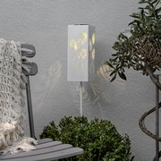 Weiße, rechteckige Garten-Stehleuchte mit Blattmuster-Ausstanzungen, warm leuchtend, in Frontansicht leicht schräg von links vor grauer Wand zwischen Stuhl und Pflanze.