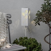 Weiße, rechteckige Garten-Stehleuchte mit Blattmuster-Ausstanzungen, warm leuchtend, in Frontansicht leicht schräg von links vor grauer Wand zwischen Stuhl und Pflanze.