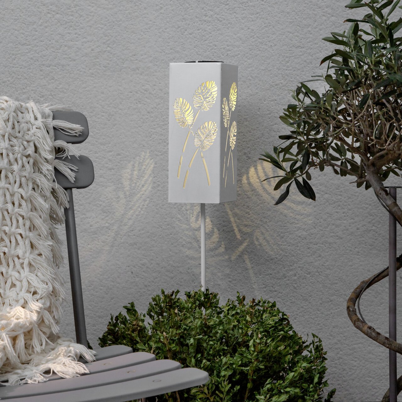 Weiße, rechteckige Garten-Stehleuchte mit Blattmuster-Ausstanzungen, warm leuchtend, in Frontansicht leicht schräg von links vor grauer Wand zwischen Stuhl und Pflanze.