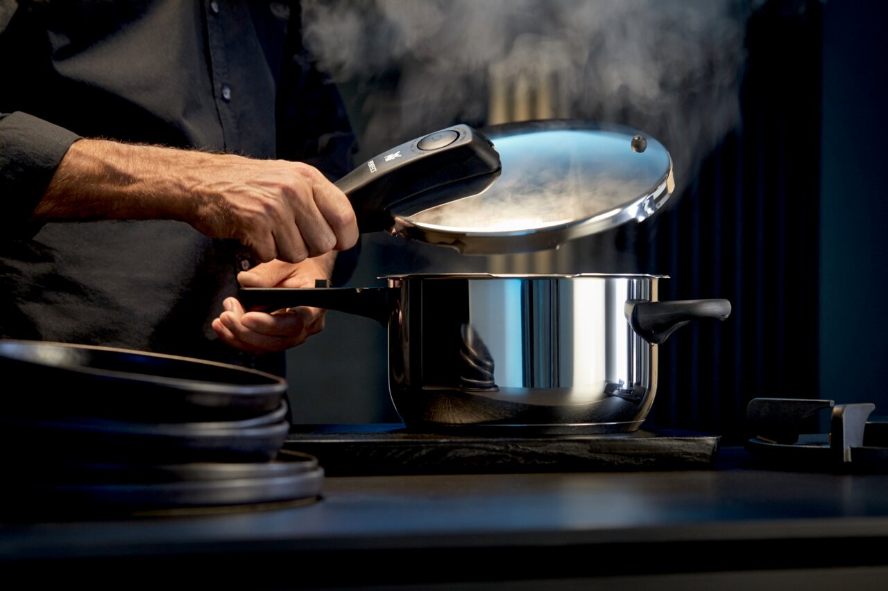 Mann öffnet einen dampfenden Schnellkochtopf aus Edelstahl in einer modernen Küche, seitliche Perspektive.