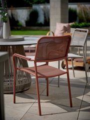 Roter Stapelsessel aus Metall mit Armlehnen, seitliche Perspektive auf einer Terrasse neben einem runden Tisch.