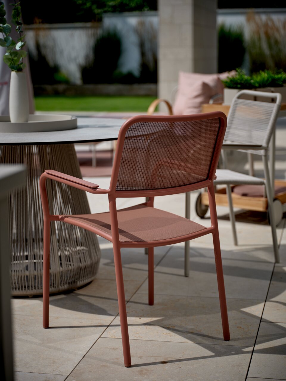 Roter Stapelsessel aus Metall mit Armlehnen, seitliche Perspektive auf einer Terrasse neben einem runden Tisch.