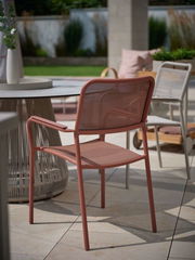 Roter Stapelsessel aus Metall mit Armlehnen, seitliche Perspektive auf einer Terrasse neben einem runden Tisch.