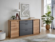 Moderne Kommode/Sideboard in Holzoptik mit anthrazitfarbenen Fronten und schwarzen Griffen, frontal aus leicht schräger Perspektive im hellen Wohnzimmer inszeniert