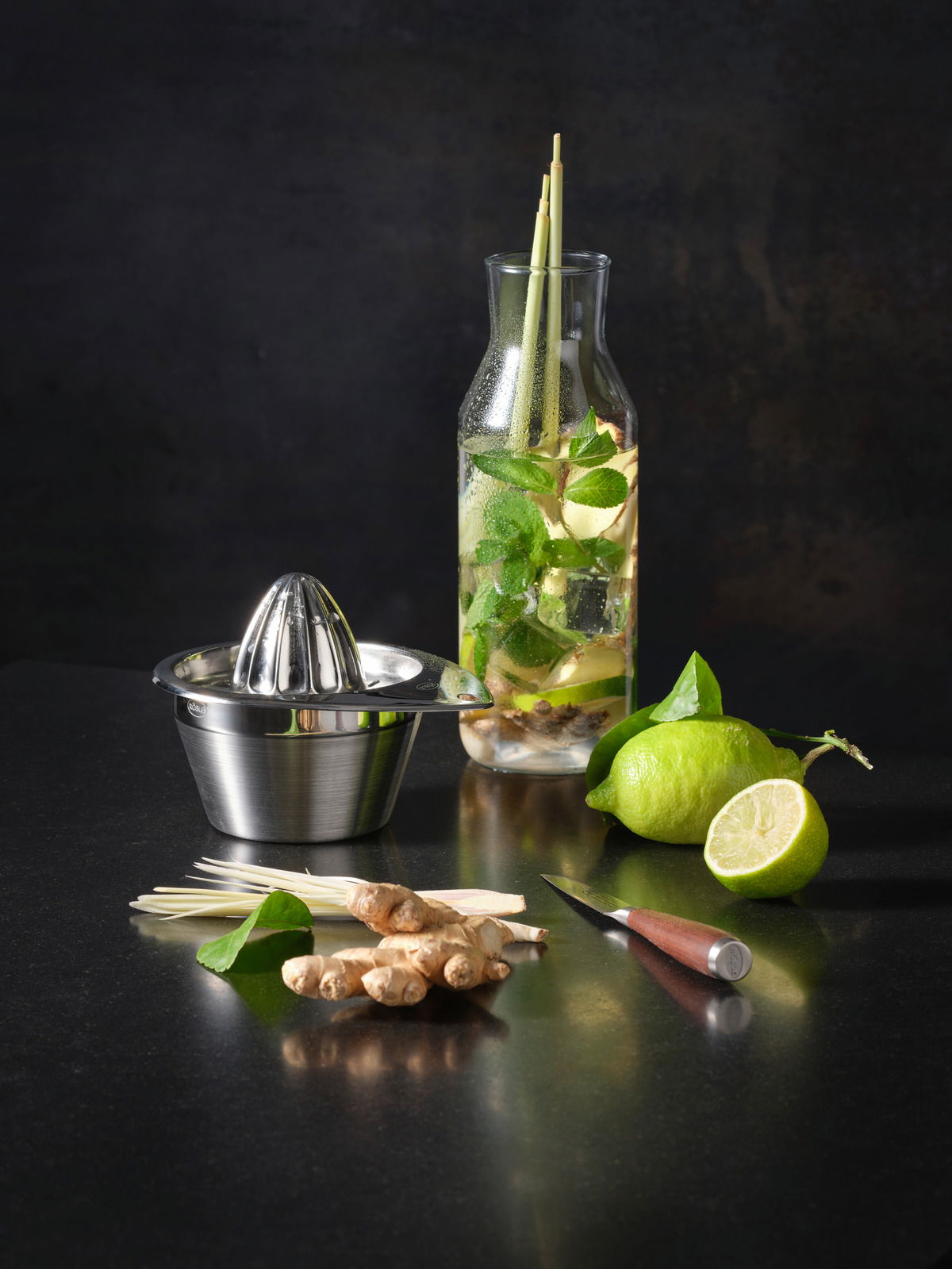 Zitronenpresse aus Edelstahl in seitlicher Perspektive, umgeben von frischen Zutaten wie Limetten, Ingwer und Zitronengras auf einer dunklen Arbeitsfläche.