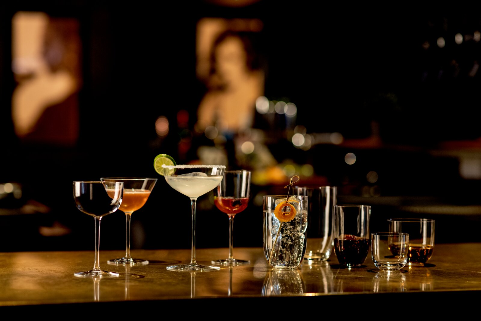 Verschiedene Cocktailgläser mit bunten Drinks und Garnituren (u. a. Margarita mit Salzrand und Limettenscheibe) auf einer Bar-Theke, aufgenommen aus frontaler Perspektive auf Augenhöhe mit unscharfem Bar-Hintergrund.