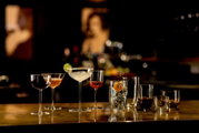 Verschiedene Cocktailgläser mit bunten Drinks und Garnituren (u. a. Margarita mit Salzrand und Limettenscheibe) auf einer Bar-Theke, aufgenommen aus frontaler Perspektive auf Augenhöhe mit unscharfem Bar-Hintergrund.