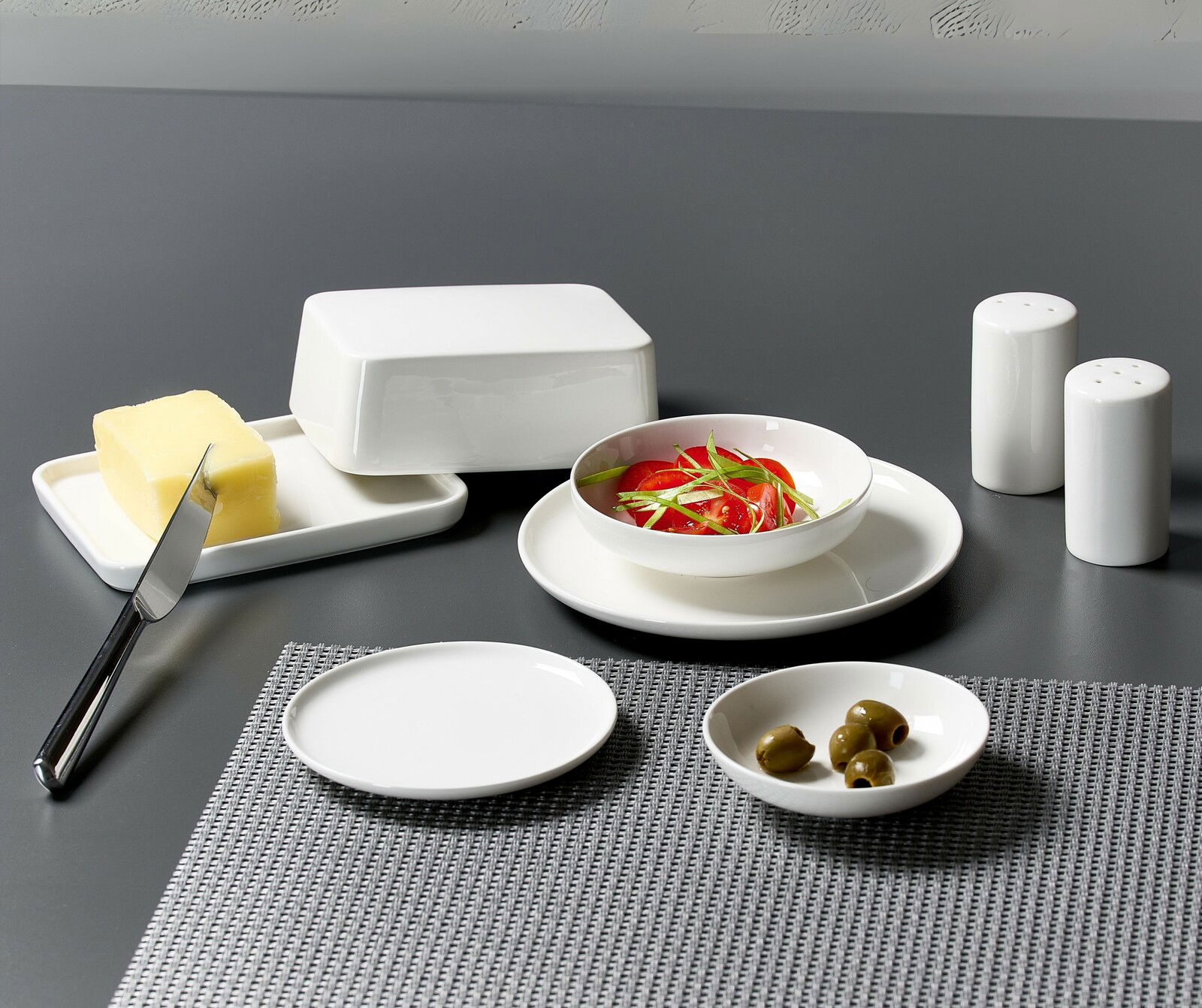 Ritzenhoff & Breker Butterdose SKAGEN Tischgedeck mit weißem Geschirrset aus der Skagen-Serie, bestehend aus einer Butterdose, einem Beilagenteller, einer Beilagenschale mit Oliven, einer Schale mit Tomatensalat und einem Salz- & Pfefferstreuerset, auf einer grauen Tischdecke von oben betrach