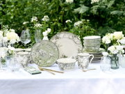 Elegantes Geschirrset mit grün-weißem Floral- und Vogelmotiv (Teller, Schalen, Tassen) auf gedecktem Gartentisch mit Gläsern und weißen Blumen, Perspektive: frontale Tischansicht auf Augenhöhe