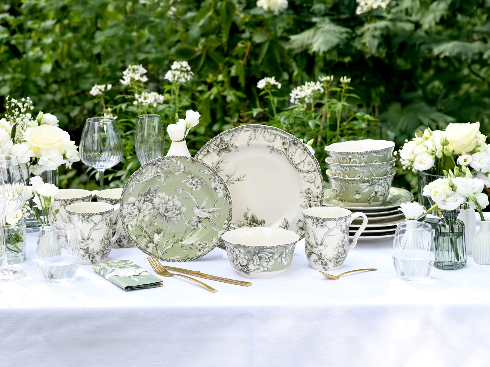 Elegantes Geschirrset mit grün-weißem Floral- und Vogelmotiv (Teller, Schalen, Tassen) auf gedecktem Gartentisch mit Gläsern und weißen Blumen, Perspektive: frontale Tischansicht auf Augenhöhe