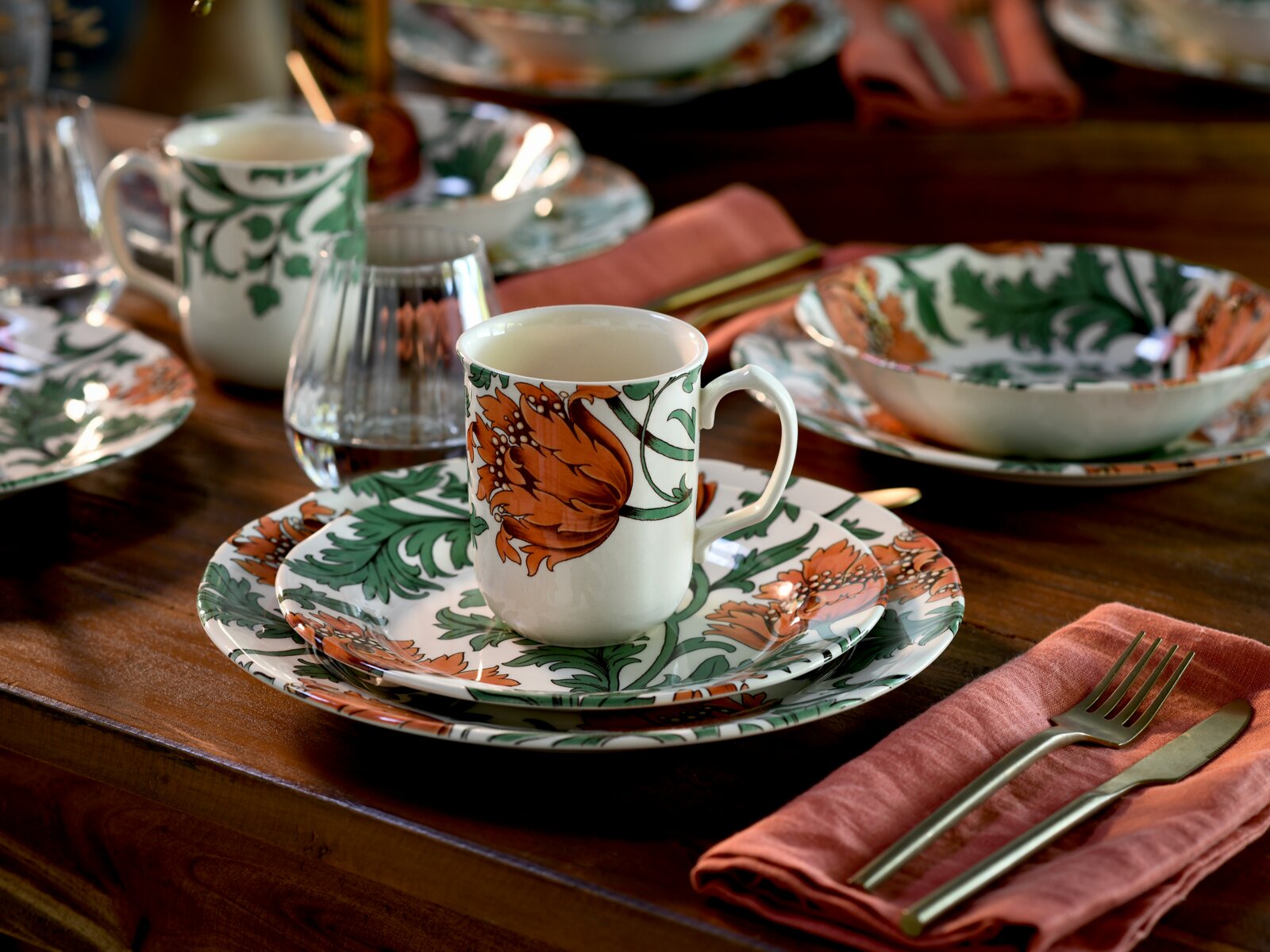 Florales Keramikgeschirr-Set mit Tasse, Teller und Schale in Grün und Orange auf gedecktem Holztisch, perspektivisch in leichter Draufsicht von schräg vorn