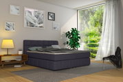Modernes Boxspringbett in einem stilvollen Schlafzimmer mit großen Fenstern und Blick auf einen grünen Garten, seitliche Perspektive.