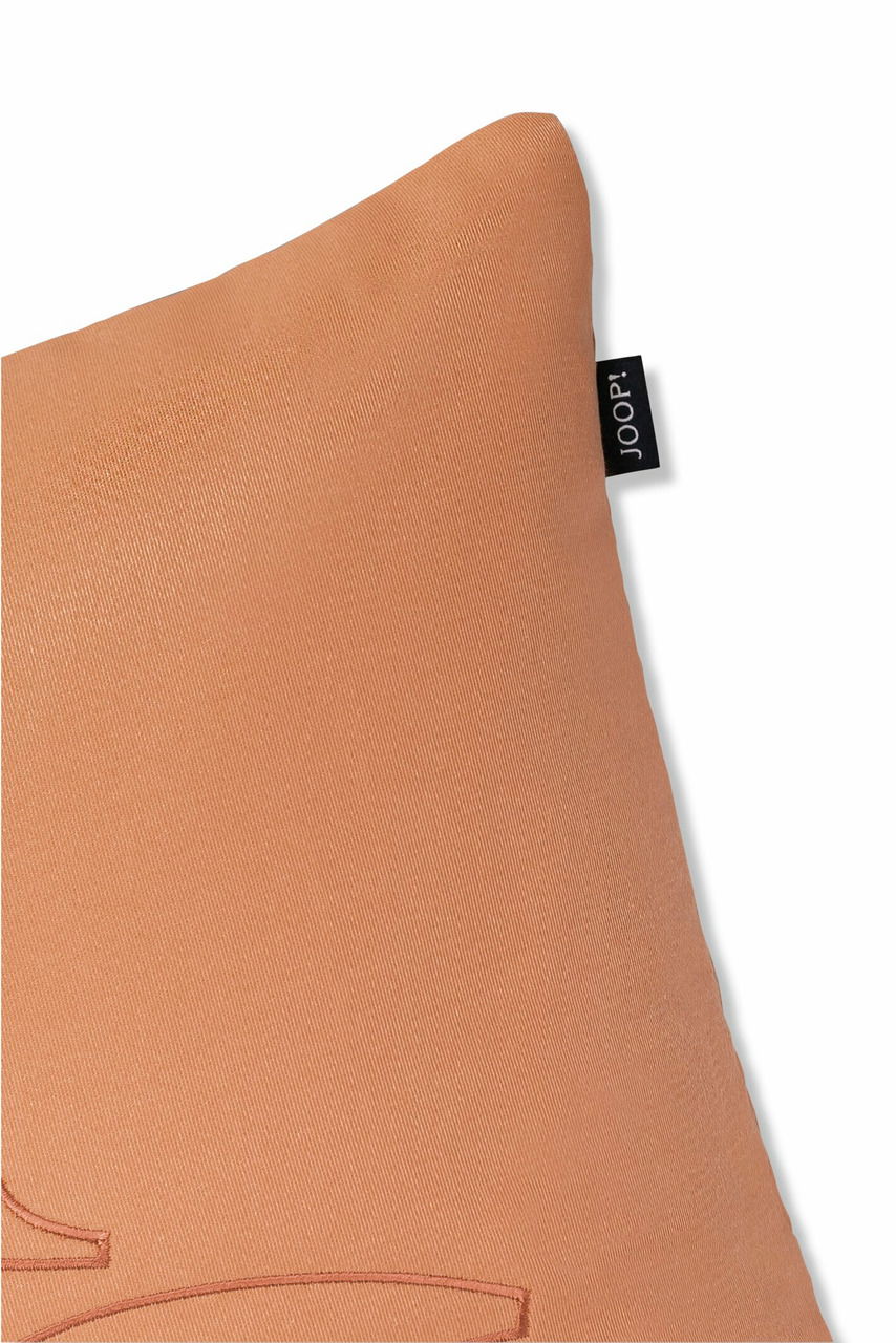 Nahaufnahme einer braunen Zierkissenhülle mit einem kleinen schwarzen Etikett an der Seite, perspektivisch von oben rechts fotografiert.