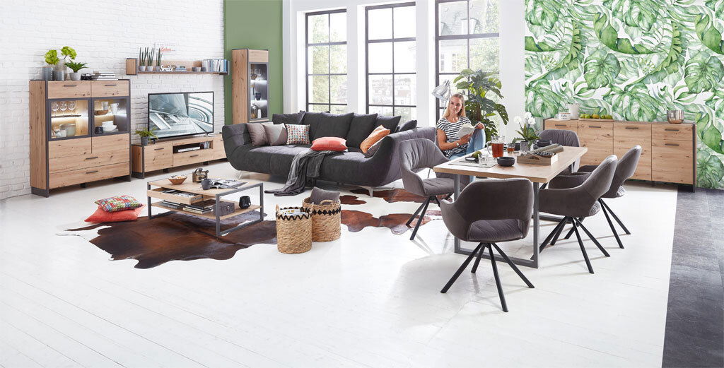 Moderne Wohnzimmermöbel in einem hellen Raum mit großen Fenstern, bestehend aus einer Vitrine, einem TV-Lowboard, einem Couchtisch und einem Esstisch mit Stühlen, aus der Perspektive des gesamten Raumes betrachtet.