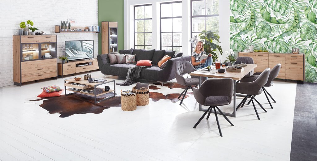 Moderne Wohnzimmermöbel in einem hellen Raum mit großen Fenstern, bestehend aus einer Vitrine, einem TV-Lowboard, einem Couchtisch und einem Esstisch mit Stühlen, aus der Perspektive des gesamten Raumes betrachtet.