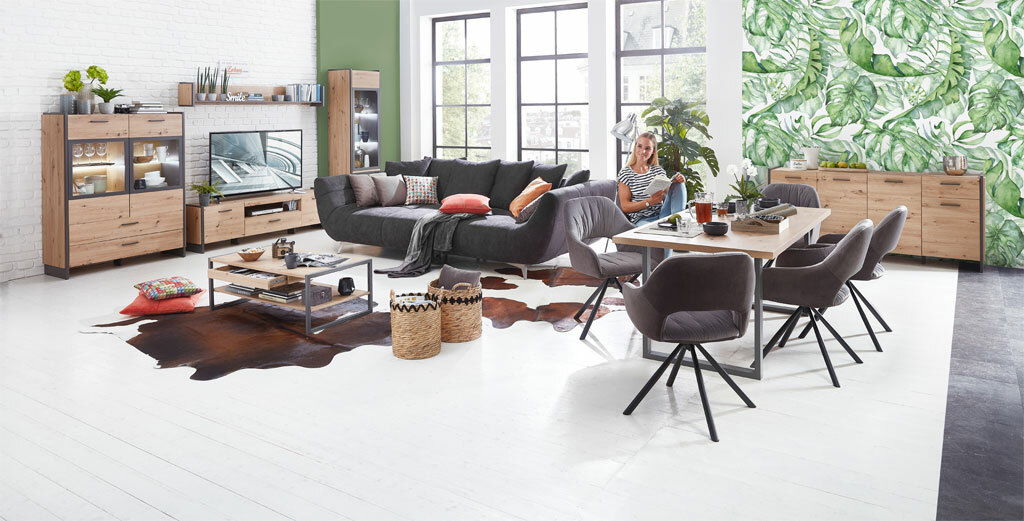 Moderne Wohnzimmermöbel in einem hellen Raum mit großen Fenstern, bestehend aus einer Vitrine, einem TV-Lowboard, einem Couchtisch und einem Esstisch mit Stühlen, aus der Perspektive des gesamten Raumes betrachtet.