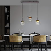 Moderne Pendelleuchte Q-KON mit drei hängenden Glaskörpern in verschiedenen Metalltönen über einem Esstisch, seitliche Perspektive.