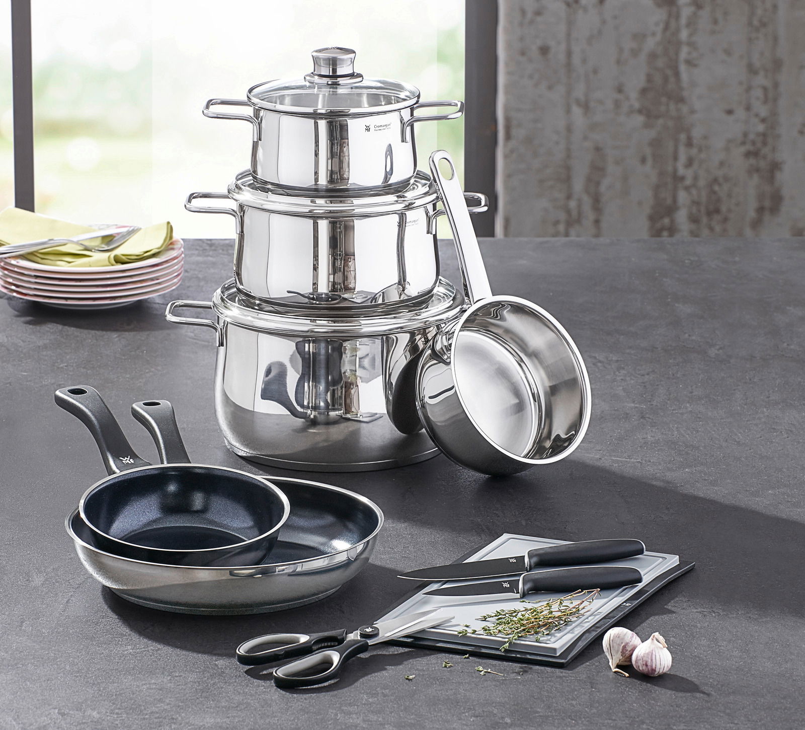 Kochtopf-Starter-Set 10-tlg. aus Edelstahl, bestehend aus mehreren Töpfen und Pfannen, mit Küchenutensilien auf einer Arbeitsfläche, seitliche Perspektive.