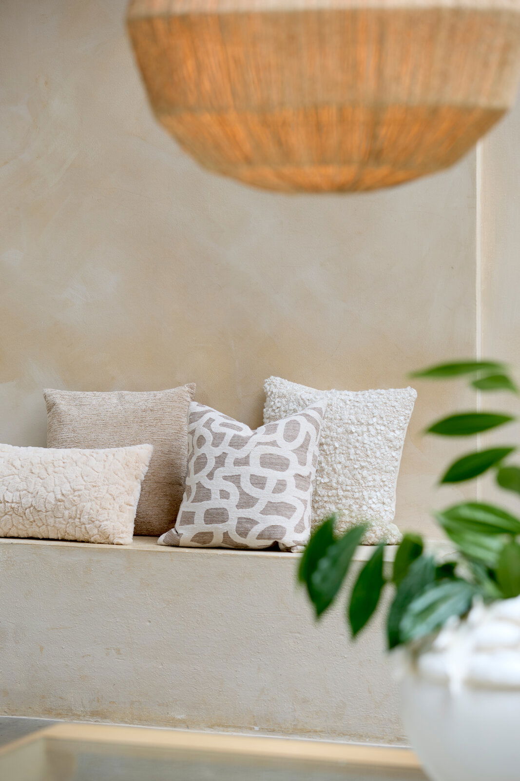 Drei dekorative Kissen in neutralen Tönen auf einer Bank, fotografiert aus einer seitlichen Perspektive. Im Vordergrund sind unscharfe grüne Blätter zu sehen.