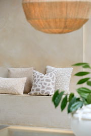 Drei dekorative Kissen in neutralen Tönen auf einer Bank, fotografiert aus einer seitlichen Perspektive. Im Vordergrund sind unscharfe grüne Blätter zu sehen.