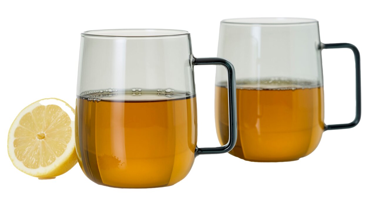 Zwei transparente Tassen mit schwarzem Griff, gefüllt mit Tee, im Vordergrund eine halbe Zitrone, seitliche Perspektive.