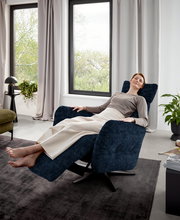 Frau entspannt in einem dunkelblauen Relaxsessel in einem modernen Wohnzimmer mit großen Fenstern, seitliche Perspektive.
