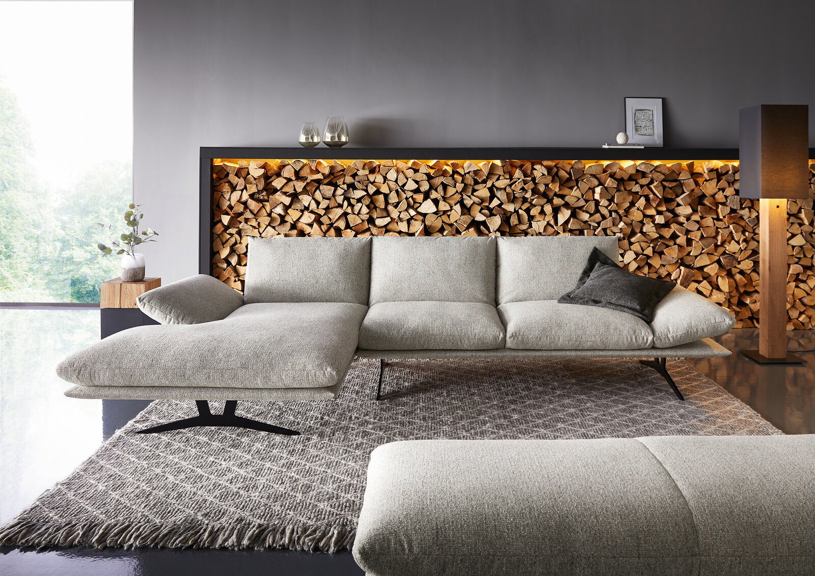 Dieter Knoll Longchairkombination FORMIA Modernes hellgraues Stoffsofa mit linksseitiger Chaiselongue und schwarzen Metallfüßen auf Teppich vor dekorativer Holzscheitwand mit LED-Licht, Frontansicht in leicht schräger Perspektive.