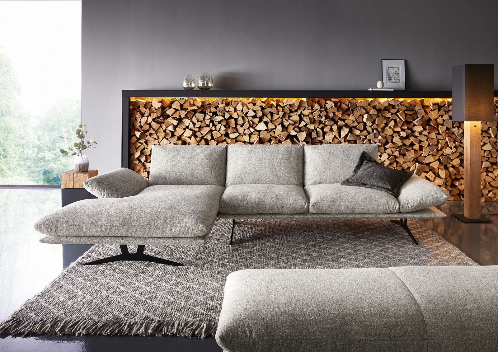 Modernes hellgraues Stoffsofa mit linksseitiger Chaiselongue und schwarzen Metallfüßen auf Teppich vor dekorativer Holzscheitwand mit LED-Licht, Frontansicht in leicht schräger Perspektive.